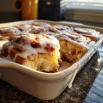 A close-up of a freshly baked cinnamon roll casserole in a white dish, generously topped with white icing.