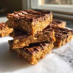 A stack of homemade, no-bake protein bars topped with drizzled chocolate, sitting on a marble surface.