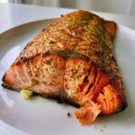 A close-up of a perfectly cooked, flaky air fryer salmon fillet seasoned with pepper and lemon zest on a white plate.