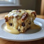 A square slice of raisin bread pudding generously topped with rich, flowing vanilla custard sauce.
