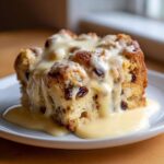 A close-up of a square slice of raisin bread pudding generously covered in vanilla cream sauce.