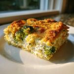 A close-up slice of amazing broccoli casserole with a creamy interior and golden, crunchy topping on a white plate.