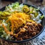A close-up of a delicious cheeseburger bowl featuring seasoned ground beef, shredded cheese, lettuce, pickles, and sauce.