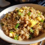 A close-up of a white bowl filled with savory chicken fried rice, featuring scrambled egg and green onions.