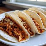 Four soft tortilla chicken tacos filled with shredded, saucy chicken and topped with white cheese.