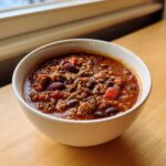 A close-up of a white bowl filled with rich, thick crockpot chili featuring ground meat and kidney beans.