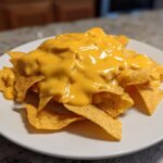 A plate of tortilla chips generously covered with smooth, bright yellow homemade nacho cheese sauce.