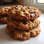 A close-up stack of three chewy chocolate chip oatmeal cookie treats on a white plate.