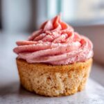 A close-up shot of a single, light-colored strawberry cupcake topped with a generous swirl of vibrant pink strawberry frosting.