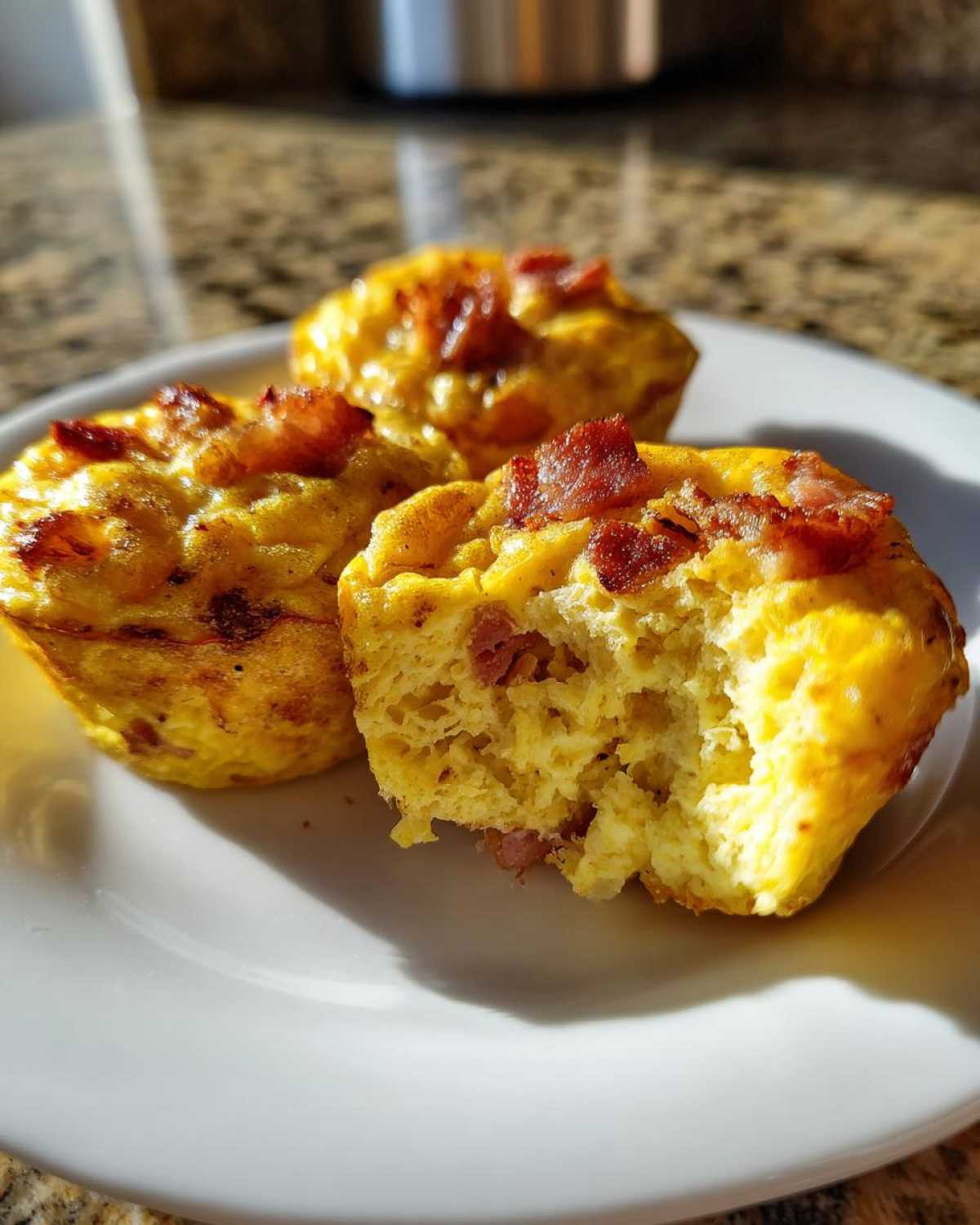 Three golden baked egg bites, one with a bite taken out, topped with crispy bacon pieces.