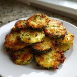 A pile of crispy, golden brown baked parmesan zucchini slices seasoned with herbs on a white plate.