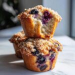 Two stacked, golden-brown blueberry muffins topped with streusel, one broken open to show the moist interior.