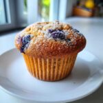 A single, golden-brown blueberry protein muffin topped with coarse sugar sits on a white plate.