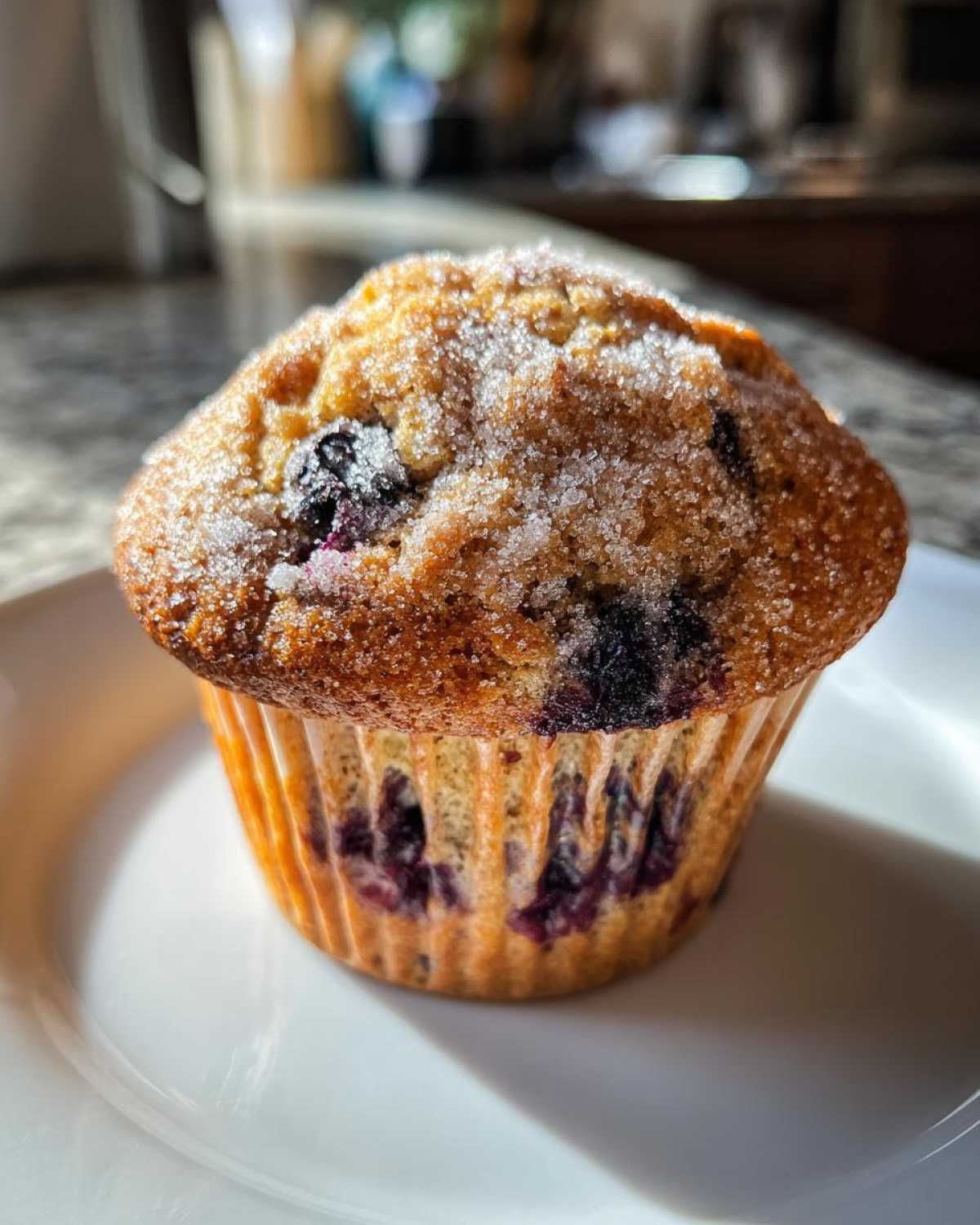 A single, golden-brown blueberry protein muffin topped generously with coarse sugar, sitting on a white plate.