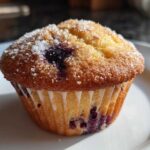 A golden brown blueberry protein muffin topped generously with coarse sugar crystals, sitting on a white plate.