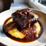 A single, dark, rich braised short ribs bone-in, resting on creamy yellow polenta and covered in thick gravy.
