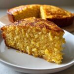A close-up of a moist, golden slice of buttermilk cornbread served on a white plate, with the rest of the round cornbread visible in the background.
