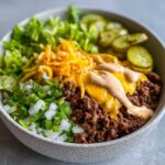 A close-up view of a cheeseburger bowl featuring seasoned ground beef, melted cheese, secret sauce, lettuce, onions, and pickles.