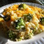 A spoonful of creamy, cheesy broccoli rice lifted from a white plate, showing melted cheddar cheese.