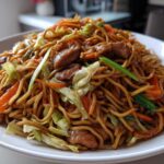 A close-up of a large serving of homemade chicken chow mein recipe with noodles, chicken pieces, cabbage, and carrots.