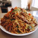 A large mound of savory chicken chow mein recipe noodles mixed with sliced beef, cabbage, and carrots on a white plate.
