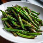 A close-up of vibrant, glossy chinese green beans with garlic sauce piled high on a white plate.