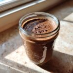 A close-up of rich chocolate overnight oats in a mason jar, topped with foamy bubbles, sitting in sunlight.