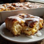 A close-up of a soft, gooey piece of cinnamon roll casserole topped with white icing, with the rest of the pan in the background.