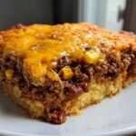 A close-up slice of cowboy cornbread casserole featuring a layer of seasoned ground beef, corn, and beans topped with melted cheddar cheese.