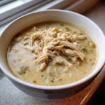 A close-up of a white bowl filled with creamy crockpot white chicken chili, topped with shredded chicken.