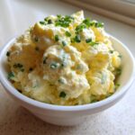 A close-up of a creamy, yellow egg salad recipe topped generously with freshly chopped green chives in a small white bowl.