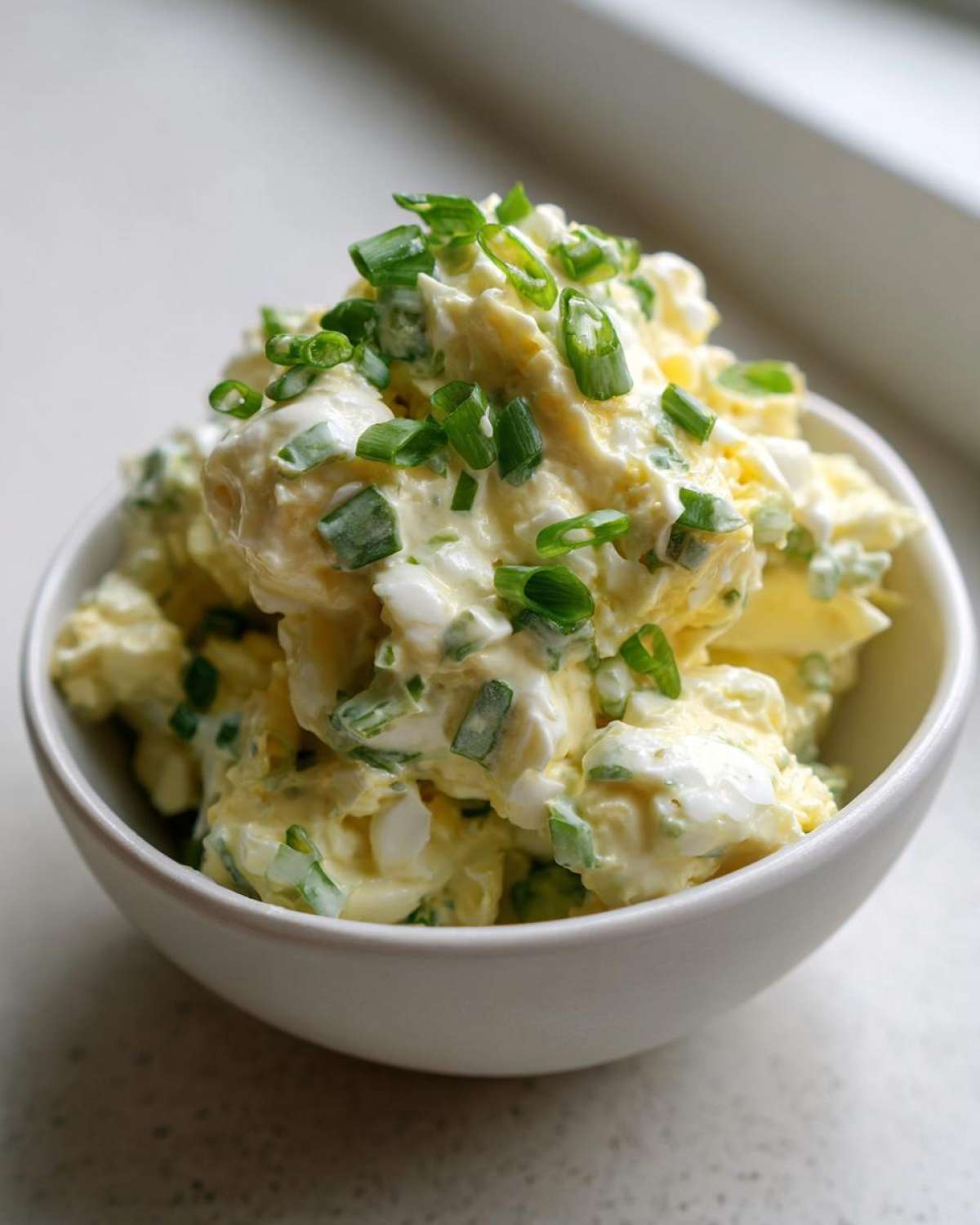A close-up of a creamy egg salad recipe served in a white bowl, generously topped with fresh chopped green onions.