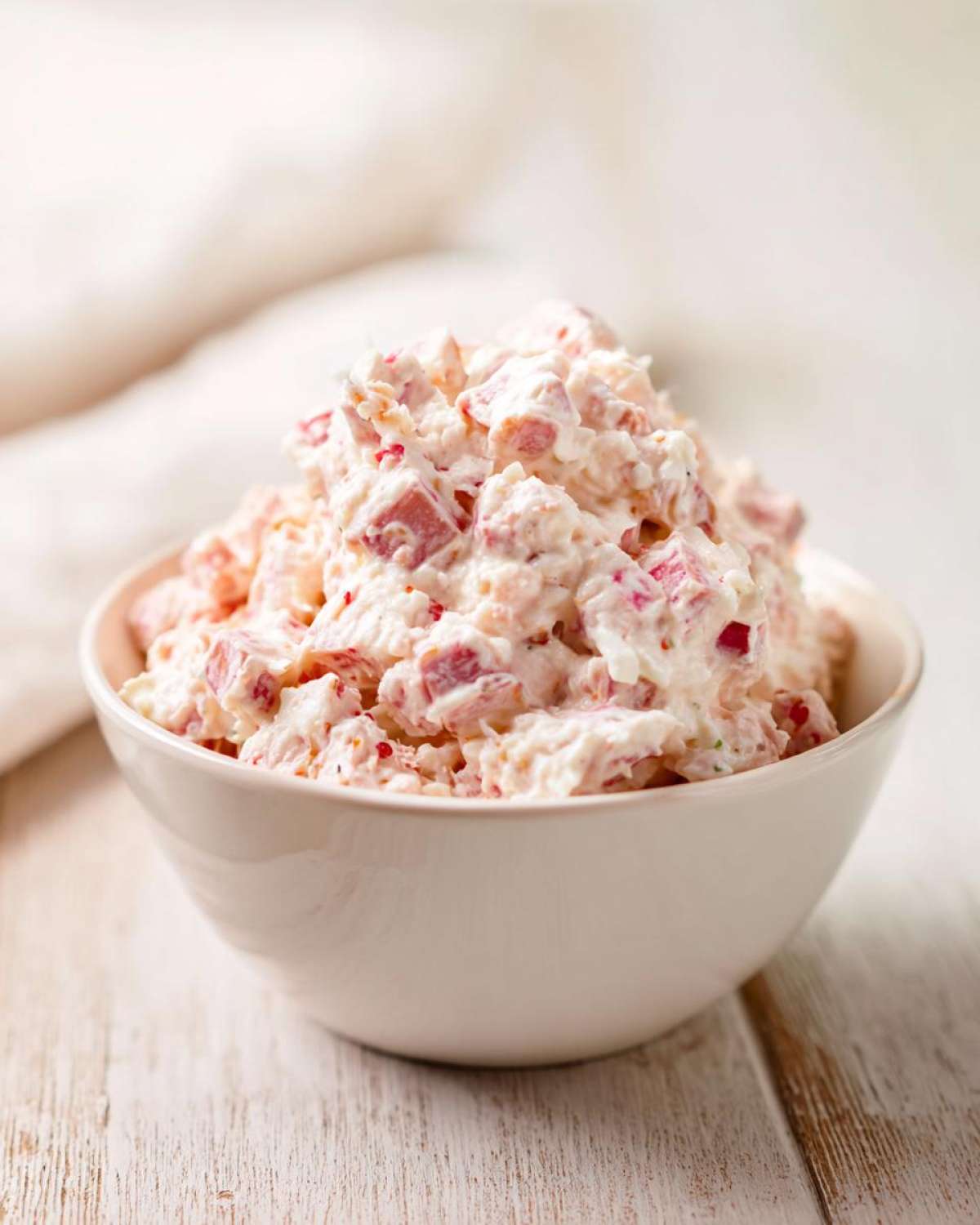 A close-up of creamy ham salad with visible chunks of pink ham served in a small white bowl on a rustic white wooden surface.