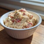 A white bowl filled with creamy ham salad featuring visible chunks of ham and red peppers, set on a wooden surface.
