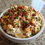 A close-up of creamy Million Dollar Dip served in a white bowl, generously topped with crumbled bacon and fresh green chives.