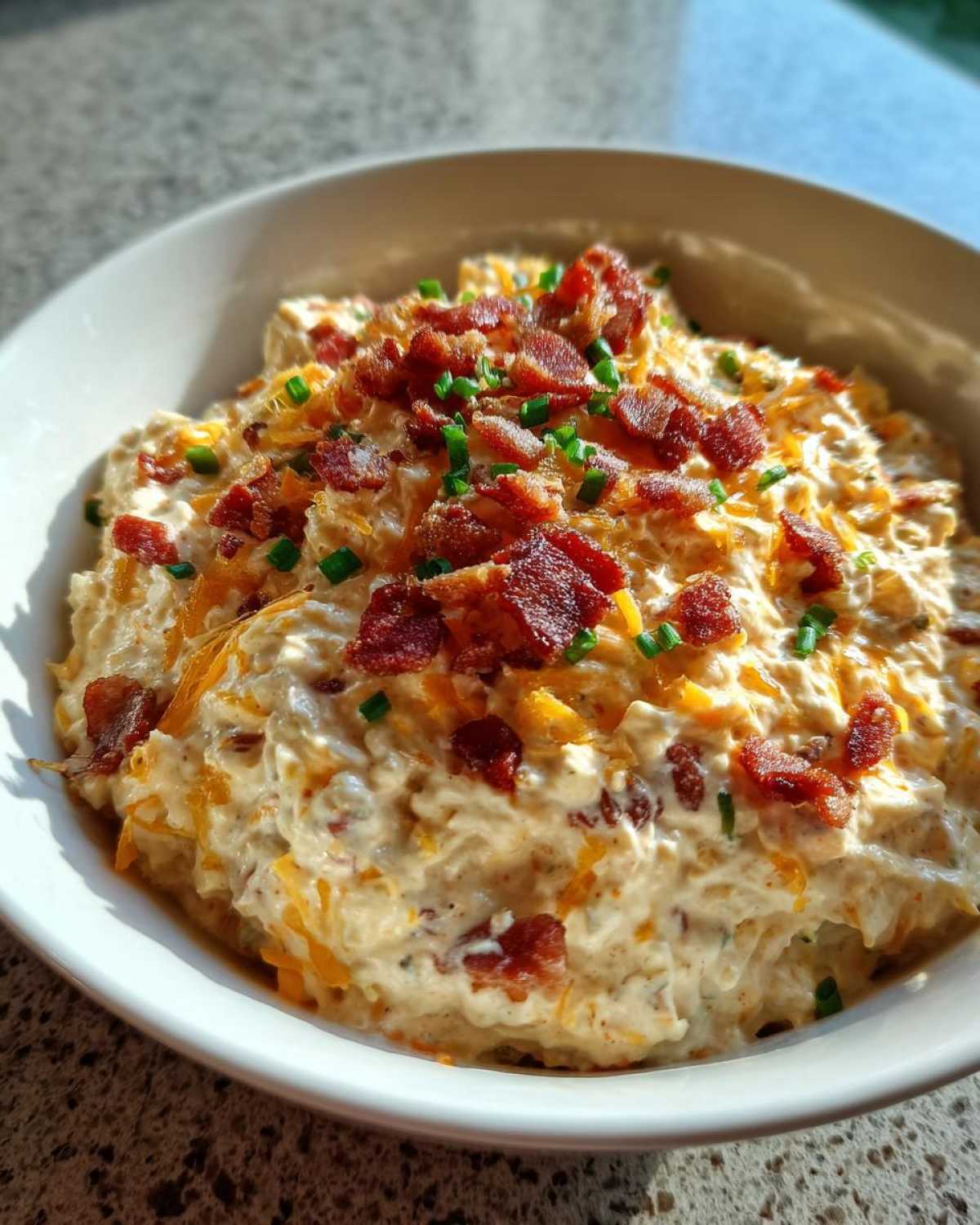 A close-up of creamy Million Dollar Dip topped generously with crumbled bacon and chopped green chives in a white bowl.