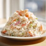 A generous mound of creamy seafood salad featuring shrimp, celery, and red onion served on a white plate.