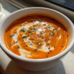 Close-up of a rich, orange tomato bisque in a white bowl, swirled with cream and topped with fresh basil.
