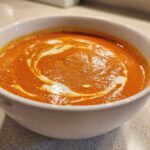 Close-up of a rich, orange tomato bisque topped with a swirl of cream in a white bowl.