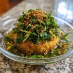 A golden brown, fried patty of crispy rice salad topped with vibrant green herbs and sesame seeds.