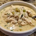 A close-up view of a hearty bowl of creamy crockpot white chicken chili, topped with shredded chicken and green chiles.