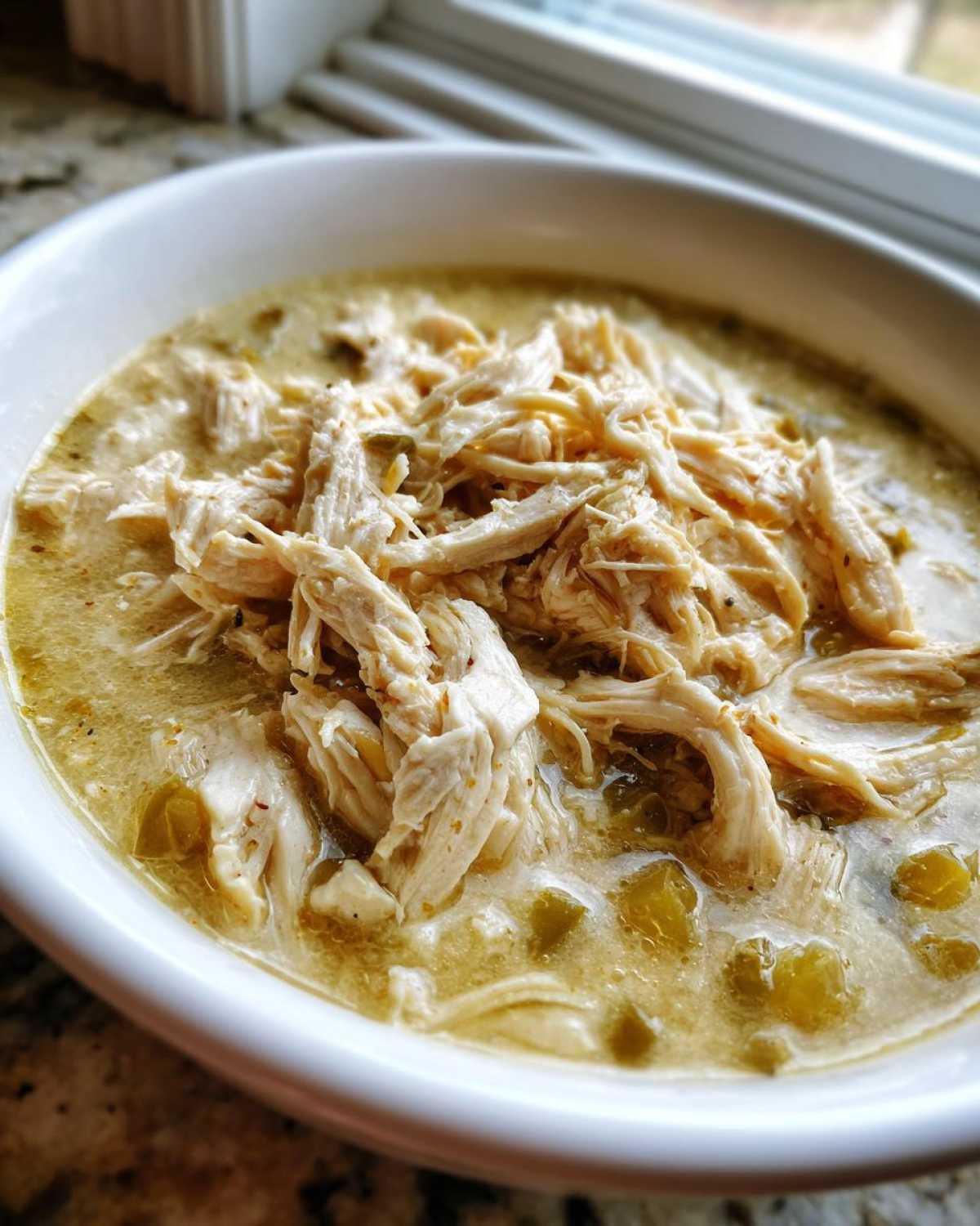 Close-up of a white bowl filled with creamy crockpot white chicken chili, topped with shredded white chicken meat and green chiles.