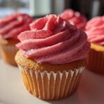 A close-up, featured image of a vanilla cupcake topped with vibrant pink strawberry frosting, with more strawberry cupcakes blurred in the background.