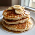 A delicious stack of three fluffy banana pancakes drizzled with syrup and topped with fresh banana slices.