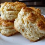 Close-up of fluffy, golden-brown buttermilk biscuits stacked on a white plate, showing their flaky interior.