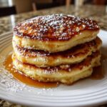 A stack of three fluffy greek yogurt pancakes drizzled with syrup and dusted with powdered sugar.