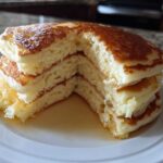 Close-up of a stack of incredibly fluffy pancakes cut to show the airy interior and golden tops, drizzled with syrup.