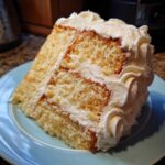 A close-up of a tall slice of moist vanilla cake recipe layered with rich white buttercream frosting.