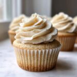 Close-up of a single, perfectly baked vanilla cupcake topped with a tall swirl of creamy, off-white vanilla frosting.