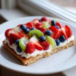 A slice of fruit pizza sugar cookie topped with cream cheese frosting, strawberries, kiwi, and blueberries.